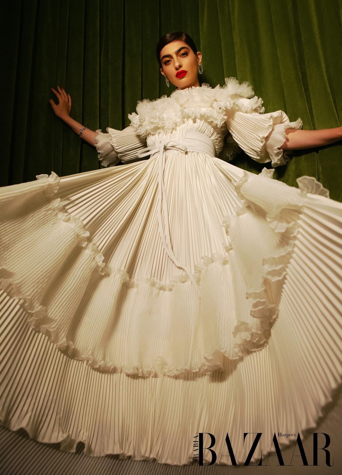 Woman wearing a white pleated gown with ruffled sleeves against a green curtain background.